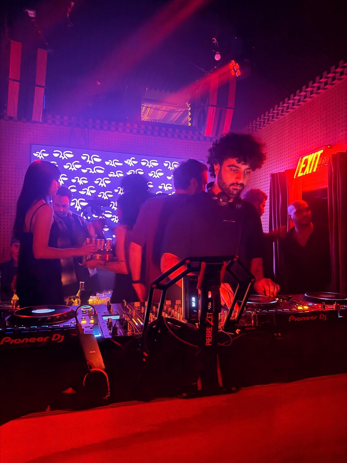 Hoodi performing at a club, red lights washing across the booth